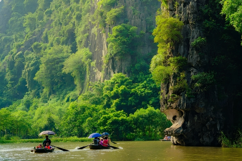 A traditional rowboat gliding smoothly through a narrow river canyon bordered by lush green limestone cliffs in Trang An, Ninh Binh.