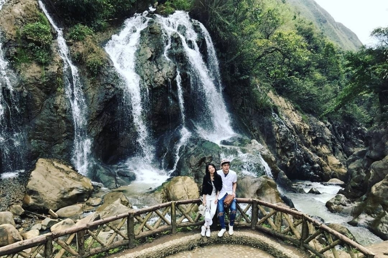A powerful white waterfall cascading down rugged stone cliffs surrounded by dense green jungle foliage in Cat Cat Village.