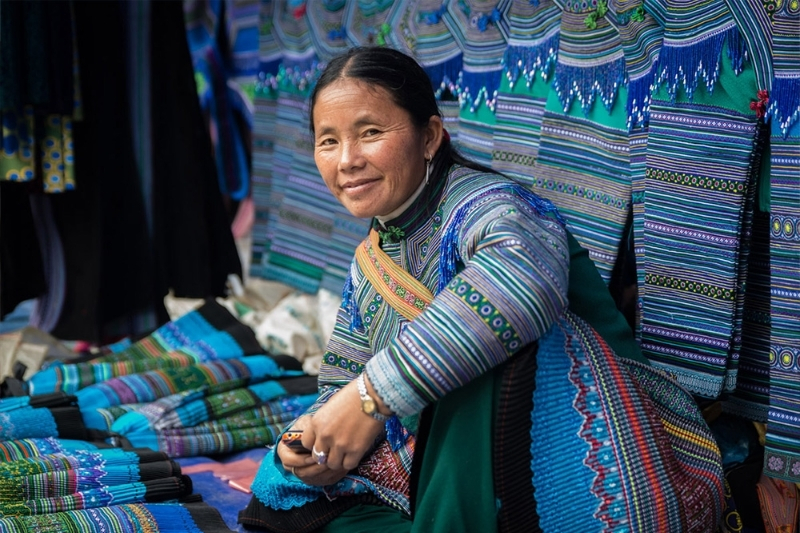 An elder Hmong woman skillfully operating a traditional wooden backstrap loom, weaving detailed geometric patterns onto hemp fabric.